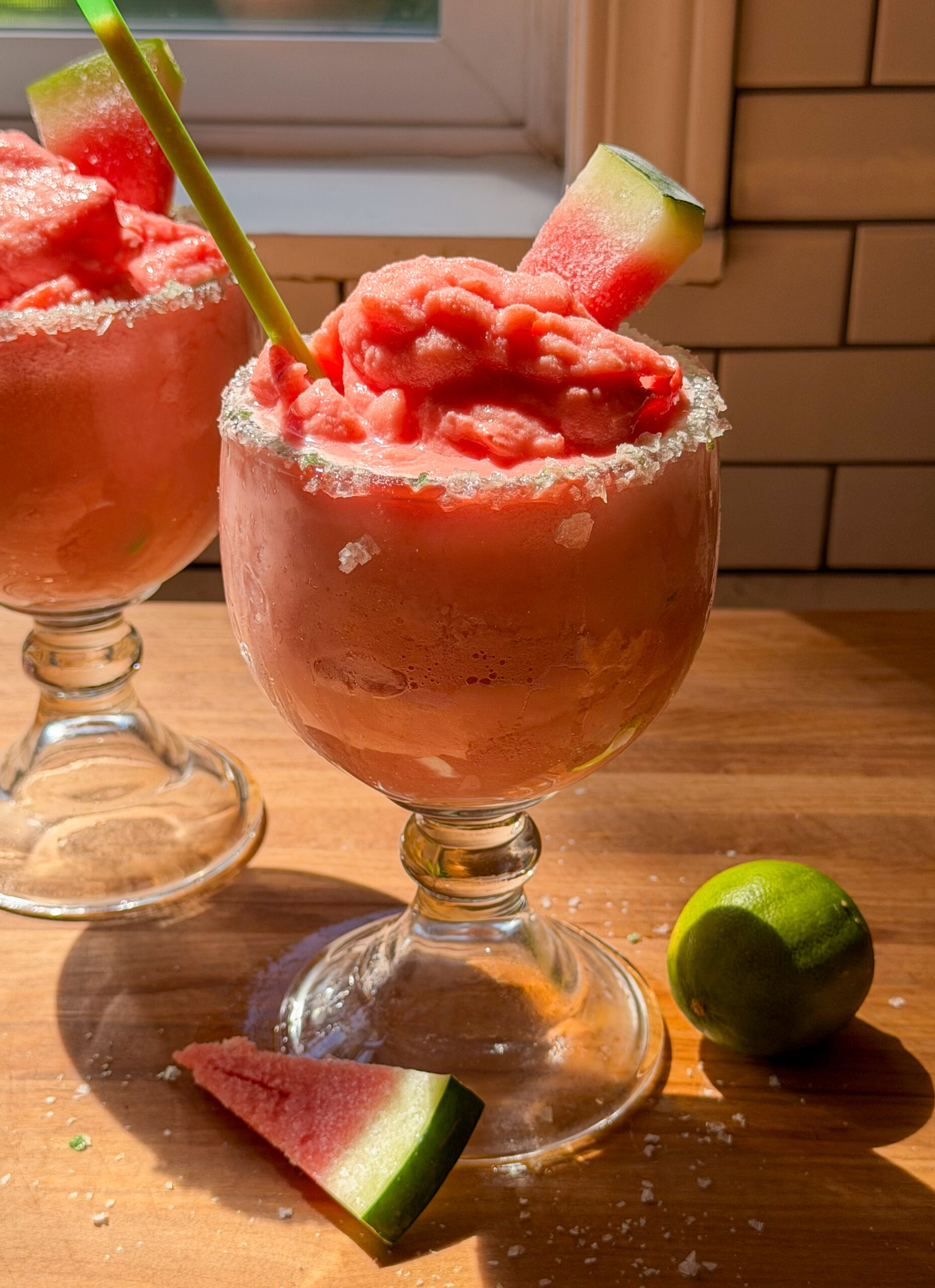 Two watermelon margarita mocktails with green salted rims on a table, garnished with watermelon wedges